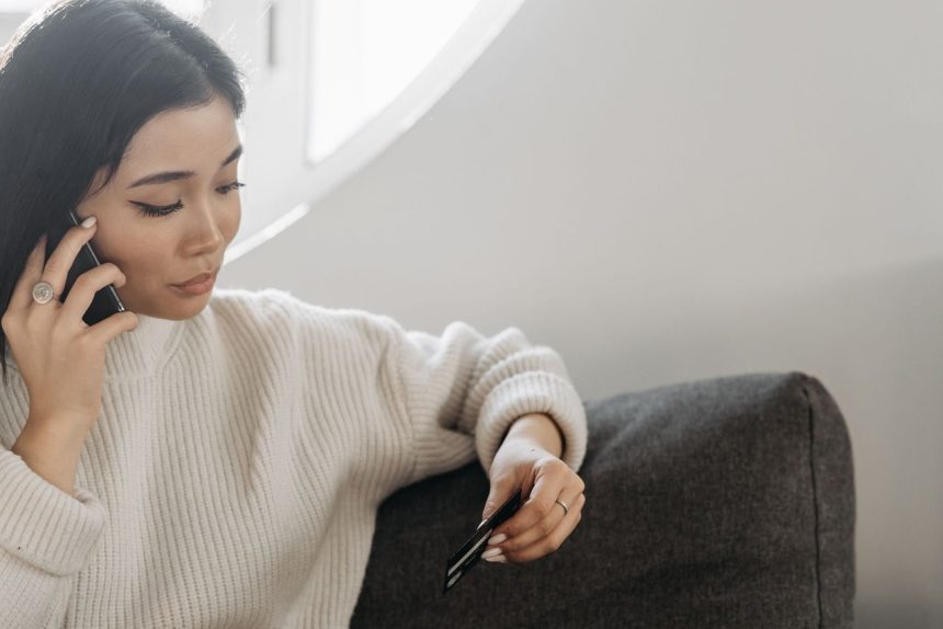 Mulher usa o celular e segura um cartão de crédito enquanto realiza uma compra online em casa.